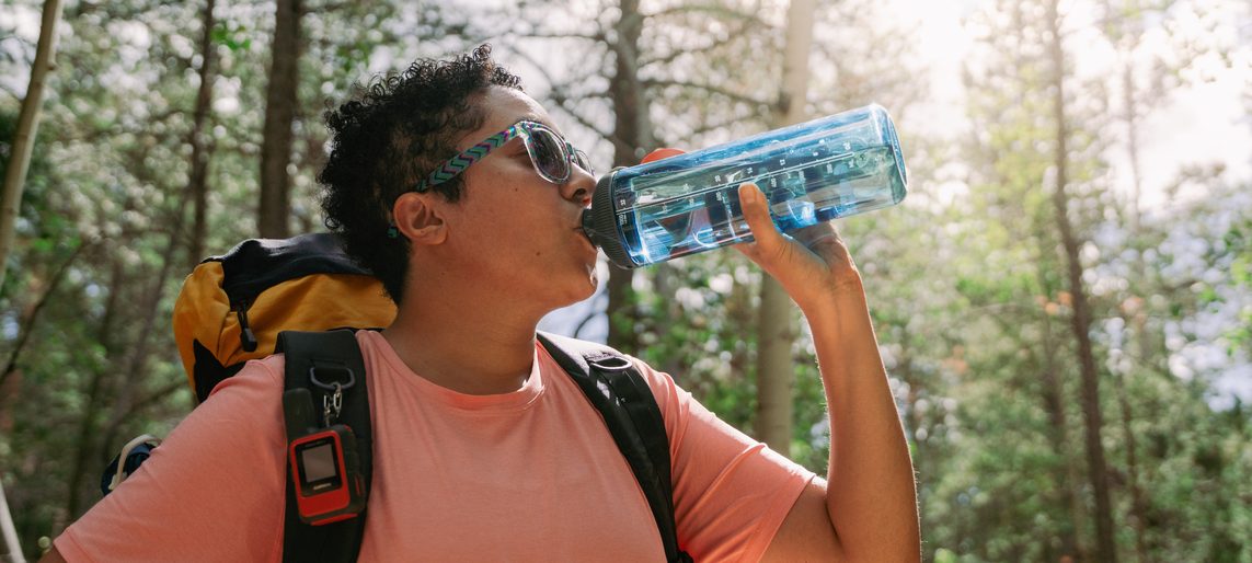 Hiker with a backpack drinking from a GoBottle held in one hand, forest behind